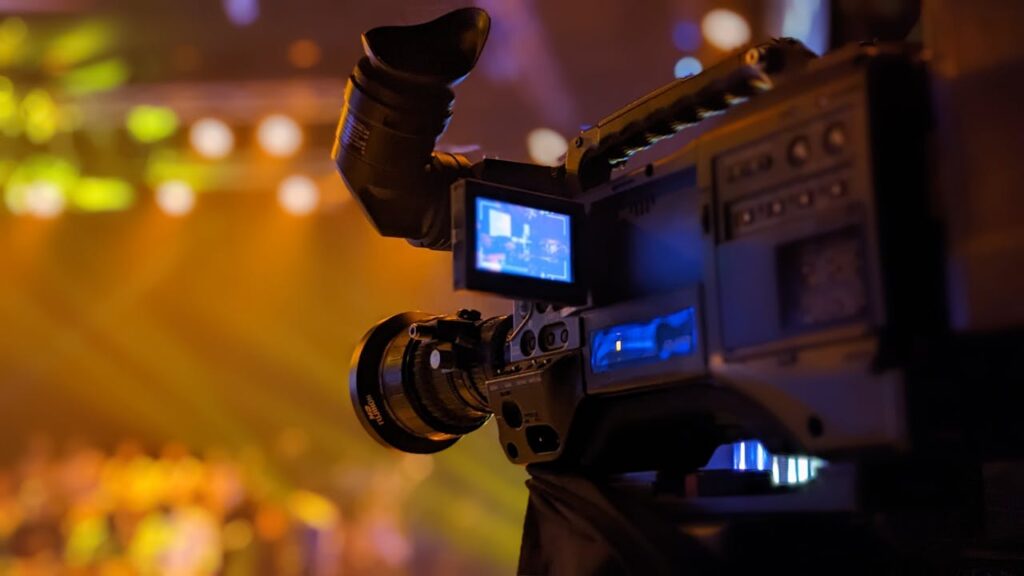 Detailed view of a video camera capturing a live event on stage with blurred lights in the background.
